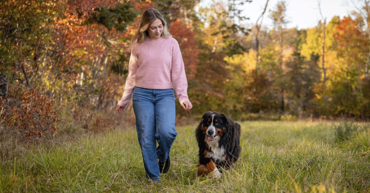 Woman walking her dog through a grassy field with autumn trees behind her