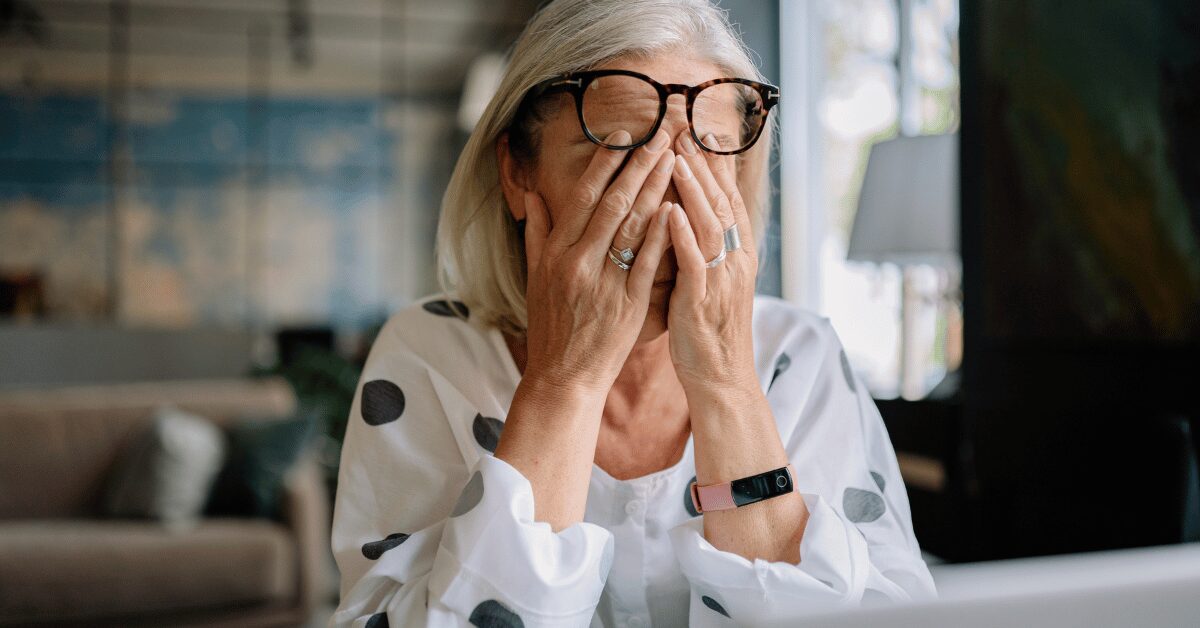 Woman in glasses pressing her hands to her face at her desk, looking overwhelmed