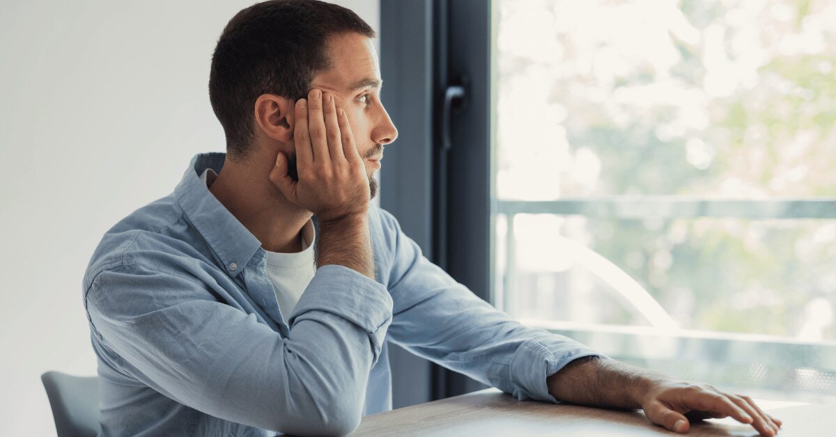 Man sitting at a table resting his head on his hand, staring out a window looking flat and unmotivated